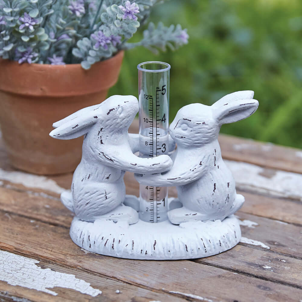 Weathered Rabbits Rain Gauge featuring two sculpted white rabbits holding a clear measuring tube in a rustic outdoor setting.