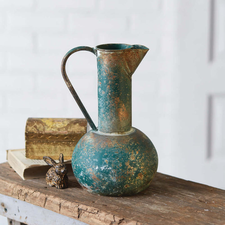 Antique oxidized metal pitcher with verdigris patina, rustic charm, and vintage appeal on a wooden surface.