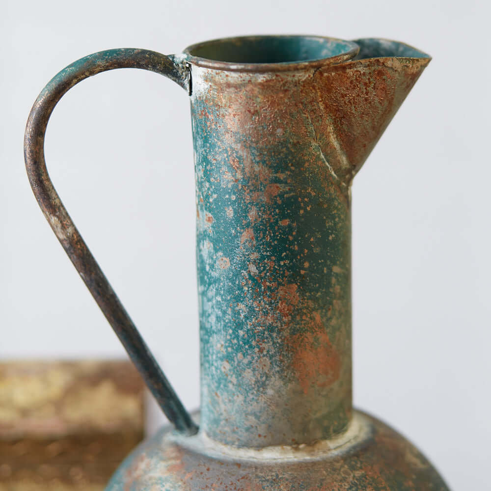 Antique oxidized metal pitcher with a weathered finish, featuring shades of blue, rust, and a vintage-inspired design.