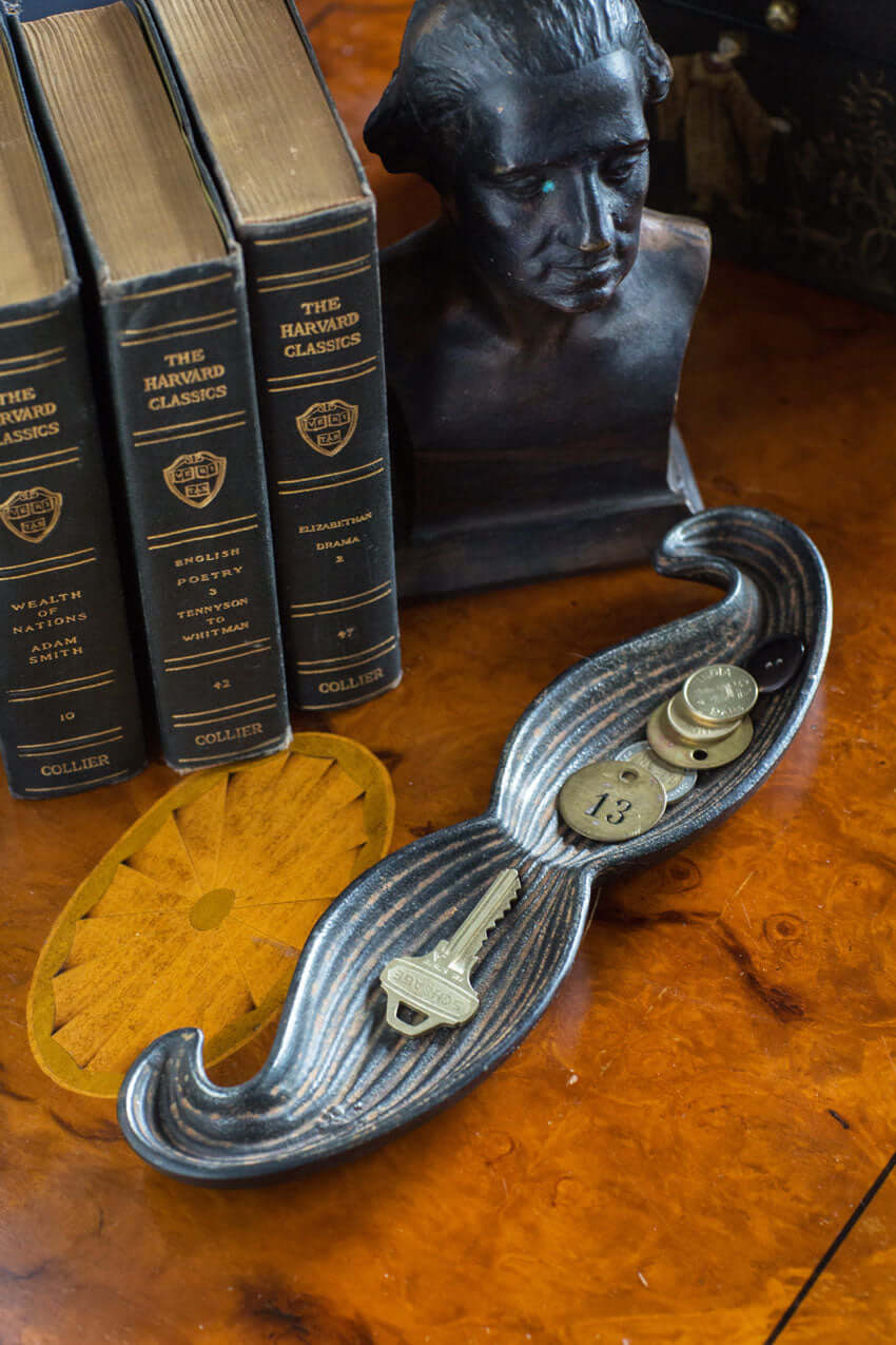 Mustache-shaped cast iron tray holding keys and coins beside vintage books and a bust sculpture, adding whimsy to decor.