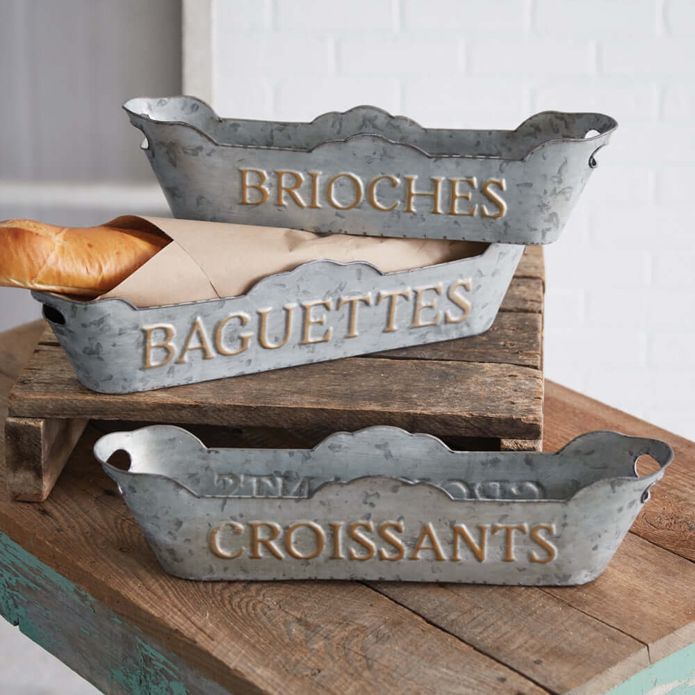 Set of 3 French country metal bread trays labeled for brioche, baguettes, and croissants on a rustic wooden surface.