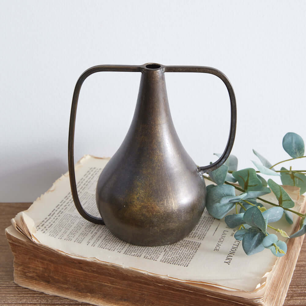 Antique bronze boho vase with dual handles on a stack of books, adorned with eucalyptus leaves.