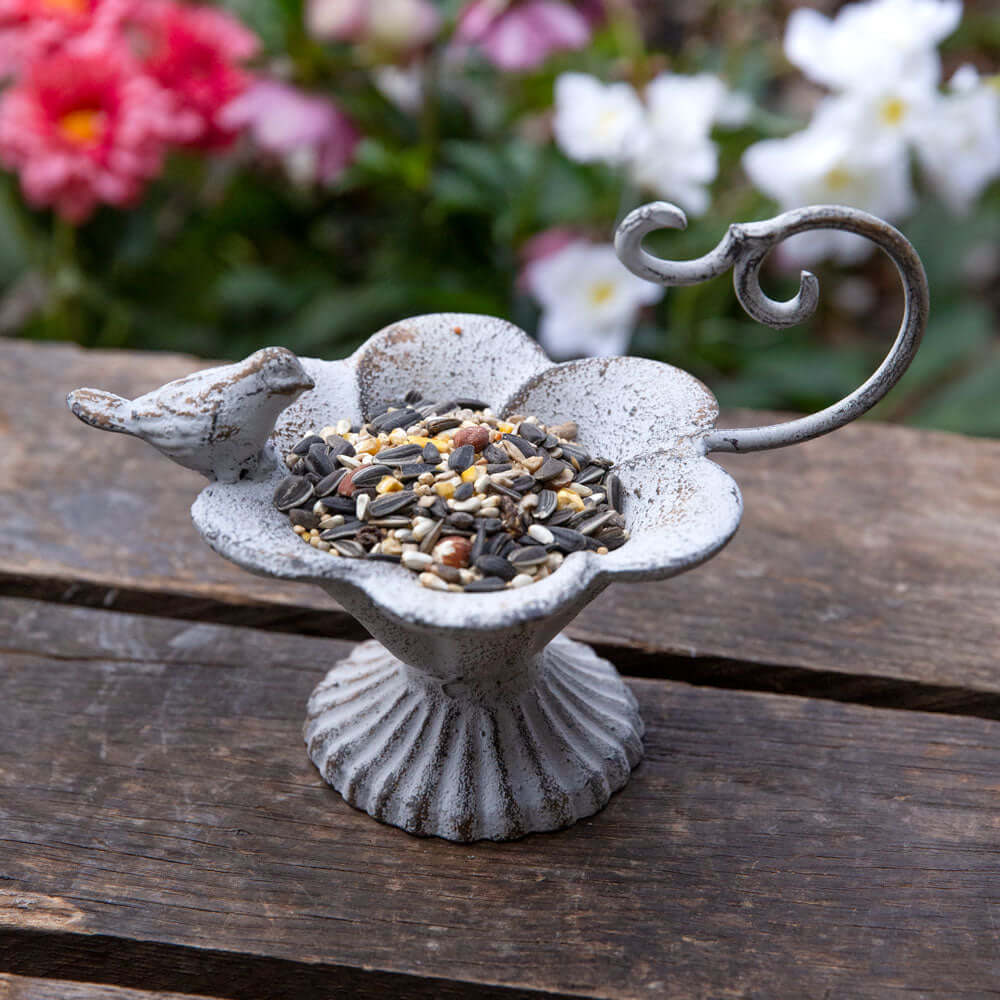 Rustic cast iron bird feeder with flower-shaped basin and bird figurine, filled with seeds, set against a colorful garden backdrop.