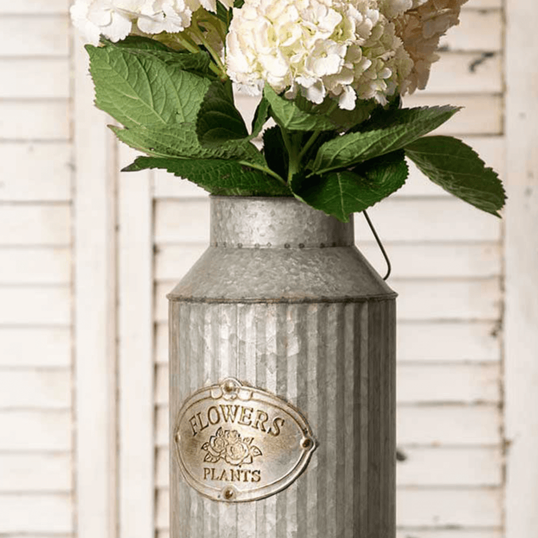 Rustic galvanized metal vase with embossed "FLOWERS PLANTS" label, filled with white floral arrangement, perfect for farmhouse décor.