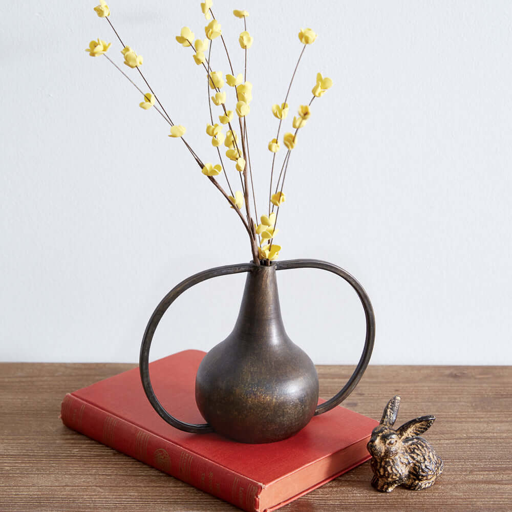 Antique bronze boho vase with yellow flowers on a red book, enhancing rustic decor and vintage charm.